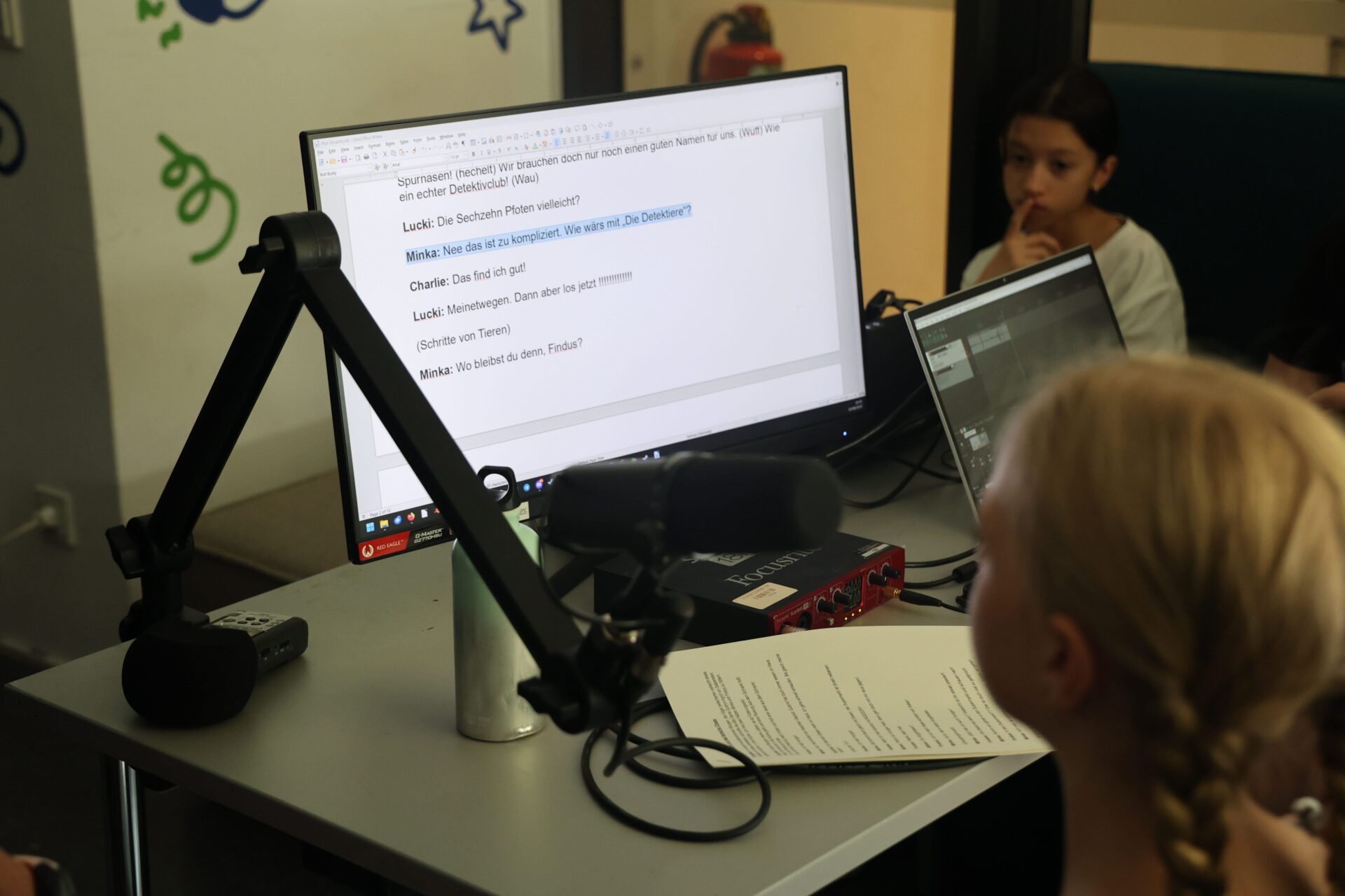 Das Bild zeigt ein Mädchen vor einem Mikrofon das einen Text von einem Computerbildschirm abliest.