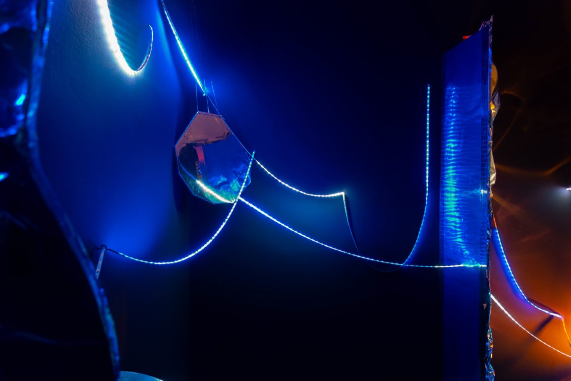 Blaue Lichtschläuche verbinden verschieden geformte Spiegel in einem dunklen Raum.