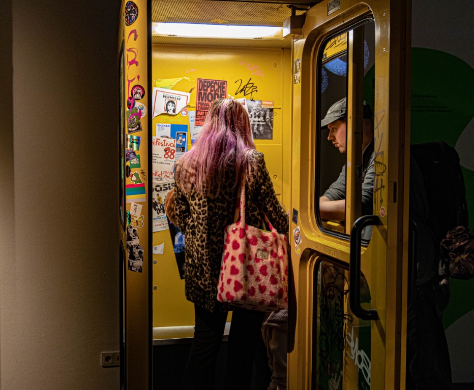 Eine Person steht in einer gelben Telefonzelle, daneben steht rechts ein Mann und sieht durch ein Fenster in die Telefonzelle hinein.