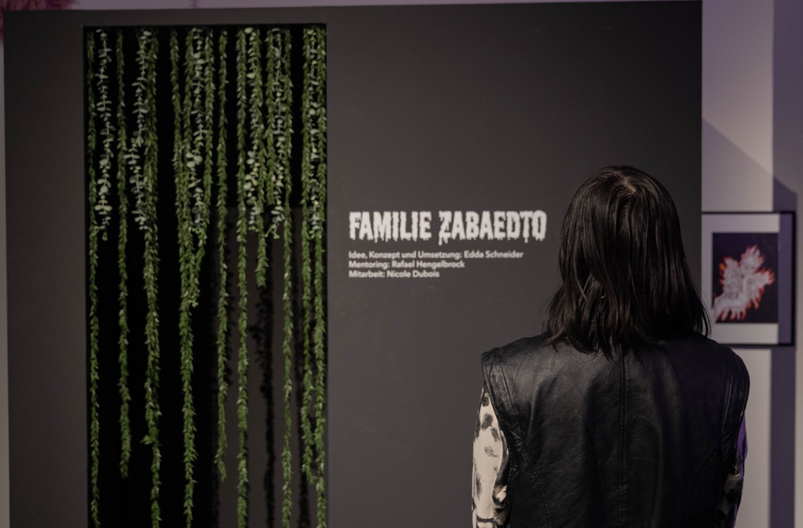 Im Vordergrund steht eine Person die auf einen kleinen schwarzen Raum schaut, dessen Eingang mit grünen, hängenden Pflanzen verziert ist und an dessen Seite der Titel 