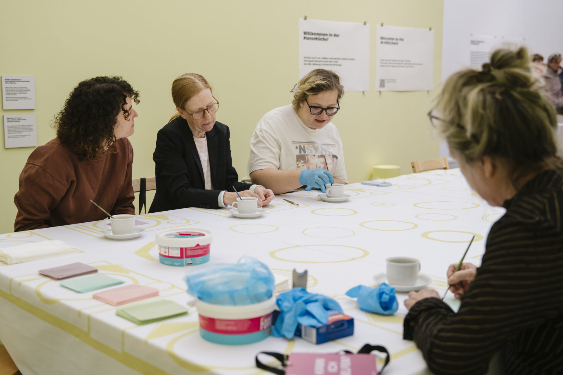 Mehrere Erwachsene sitzen an einem großen Tisch in einem Ausstellungsraum und bemalen konzentriert Tassen. Auf dem Tisch liegen Pinsel, Farben, Handschuhe und Notizzettel; im Hintergrund hängen Informationstafeln an der Wand.