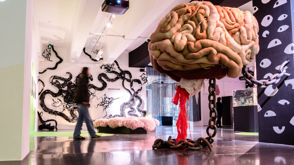 Ausstellungsansicht der Ausstellung "Wenn die Wände flüstern" auf der uzwei im Dortmunder U. Im Vordergrund die Skulptur "Veriftete Gedanken" von Lana Kattaa, das ein übergroßes Gehirn mit Ankettungen aus diversen Textilien ist.