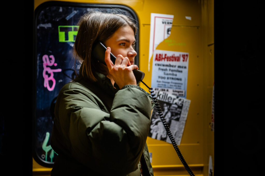 Im Vordergrund sieht man eine junge Person in einer Telefonzelle, die einen Hörer ans Ohr hält