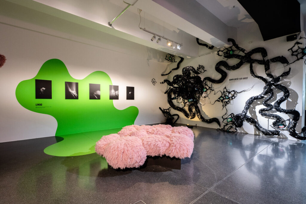 Ausstellungsansicht in die Ausstellung "Wenn die Wände Flüstern". Im Vordergrund ist eine pinke Plüsch-Sitzinsel. An der Wand links sieht man die Fotoarbeiten von Christian Plippi "Limbo", auf der rechten Wand erstreckt sich die Lichtinstallation und Skulptur "Komplexität der Angst" von Hanna Knoblauch, die schwarze Tentakeln ähnlich ist.