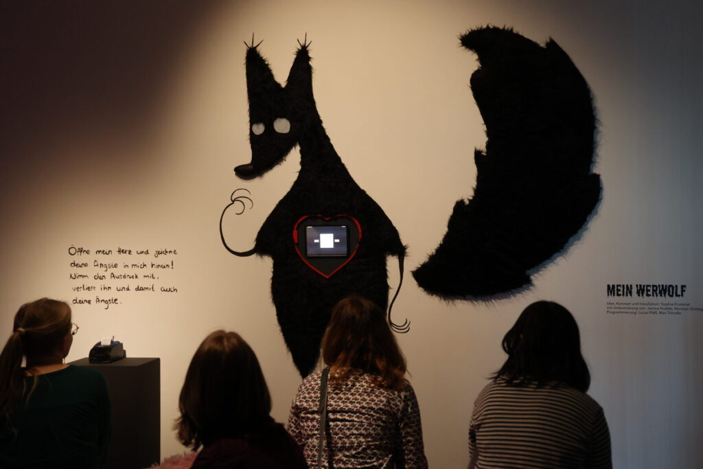 Menschen sitzen vor einer Wand, auf der die medieninstallation "Mein Werwolf" von Sophia Krutenat hängt. Es ist ein schwarzer, eindimensionaler, großer Werwolf mit einem Herz in der mitte, wo ein Monitor eingearbeitet wurde.