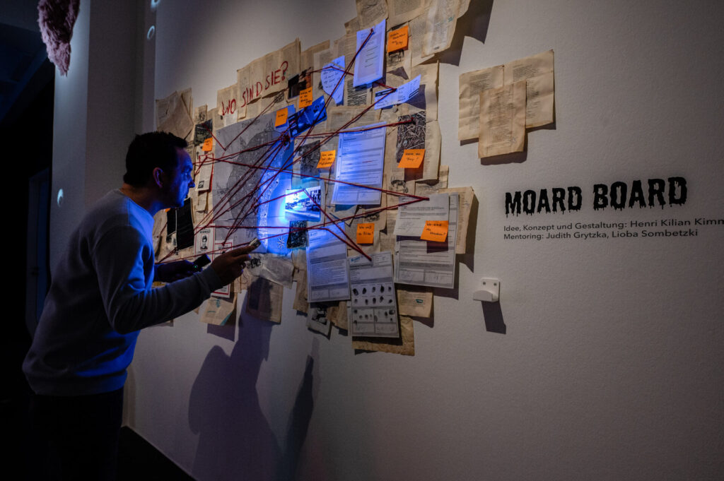 Ein Mann mit einer Blaulichttaschenlampe leuchtet eine Wand an, an der diverse Zettel hängen, die durch Stecknadeln und rotem Faden aufeinander hinweisen.