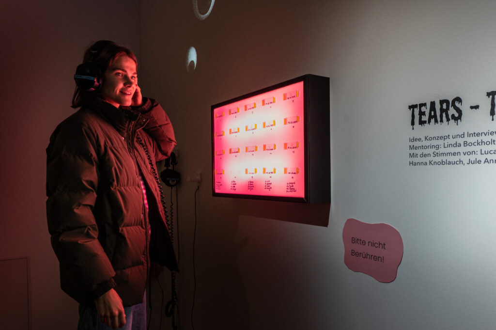 Eine Person ist links, und trägt Kopfhörer. An der Wand im rechten Bildteil ist an der Wand eiin Leuchtksten angebracht, auf dem Proben von Tränen mit Datumsangaben angeklebt sind. Das ist die Installation von Samira Szameitat in Kombination mit Audiospuren.