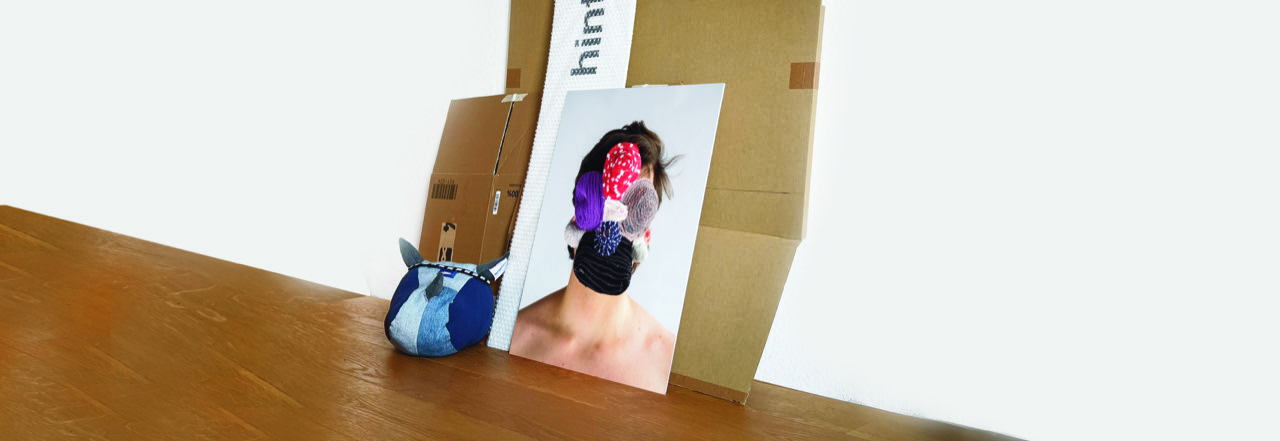 An einer Wand stehen Pappkartons und ein Porträtfoto, bei dem die/der Gezeigte eine Maske aus verschiedenfarbigen Wollknäueln trägt.