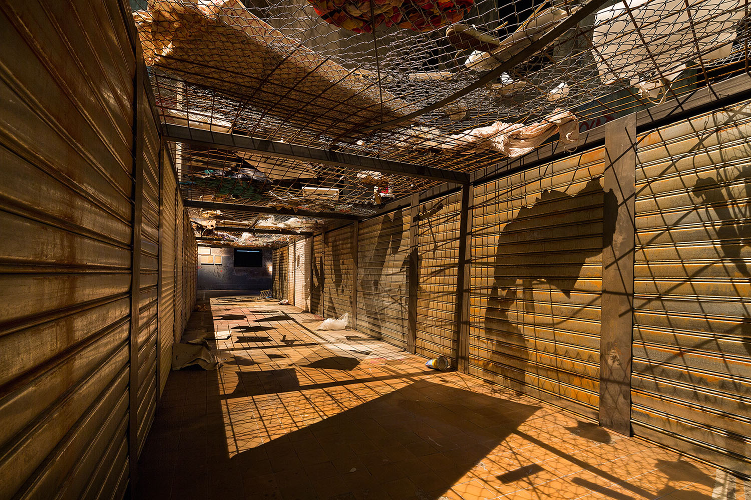 Langer, leerer Gang mit geschlossenen Metallrollläden auf beiden Seiten; über Kopf spannen sich Drahtgitter mit eingeklemmten Stoff- und Materialresten. Warmes Licht wirft harte Schatten auf den gefliesten Boden und erzeugt eine beklemmende, industriell wirkende Atmosphäre.