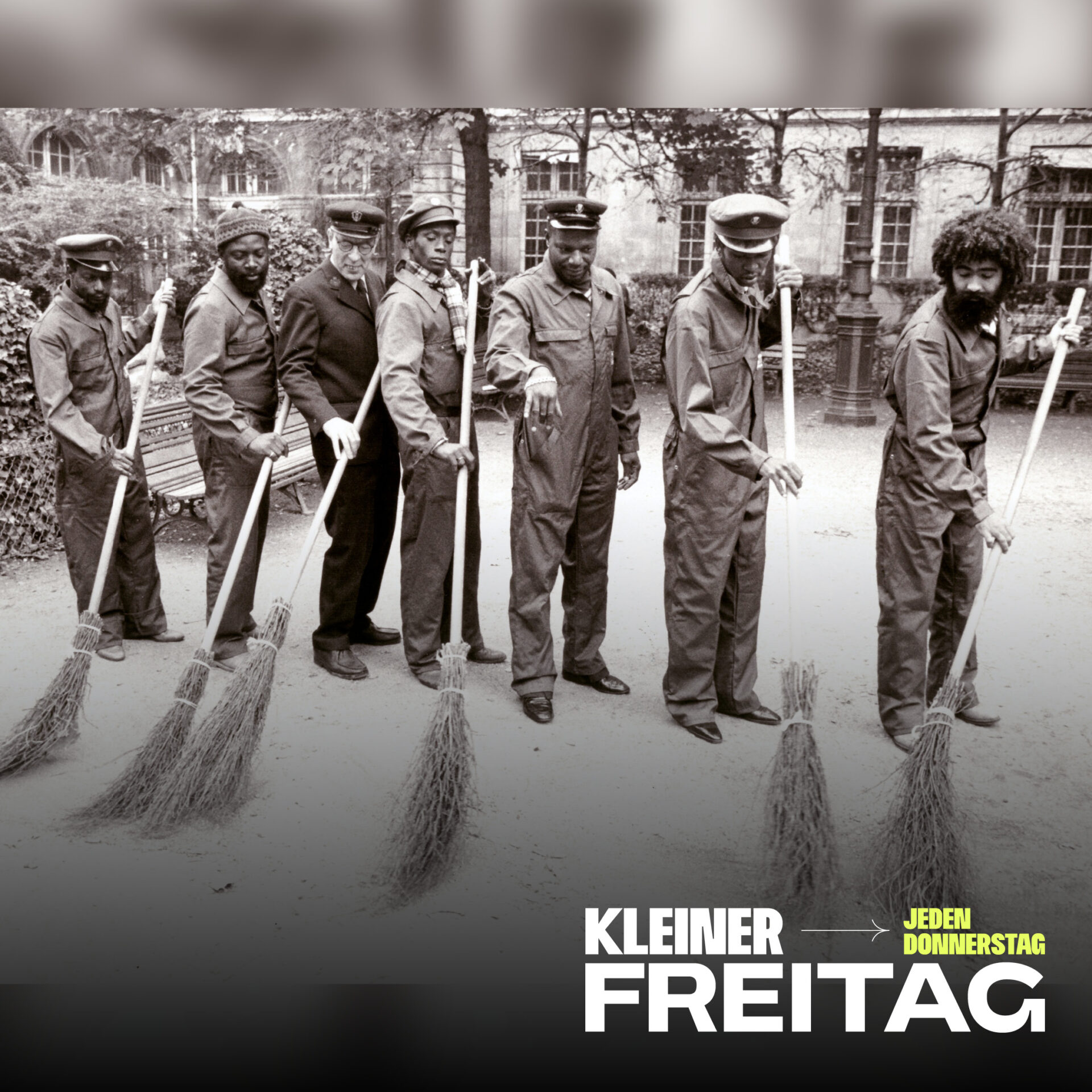 Sieben Männer in Arbeitskleidung und Mützen stehen in einer Reihe und halten Besen in einem Innenhof