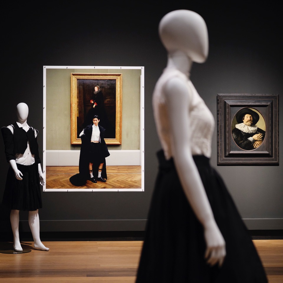 Ein Blick in die Ausstellung mit Schaufensterpuppen, die aktuelle Mode tragen, sowie einer Fotoarbeit und einem Gemälde im Hintergrund.