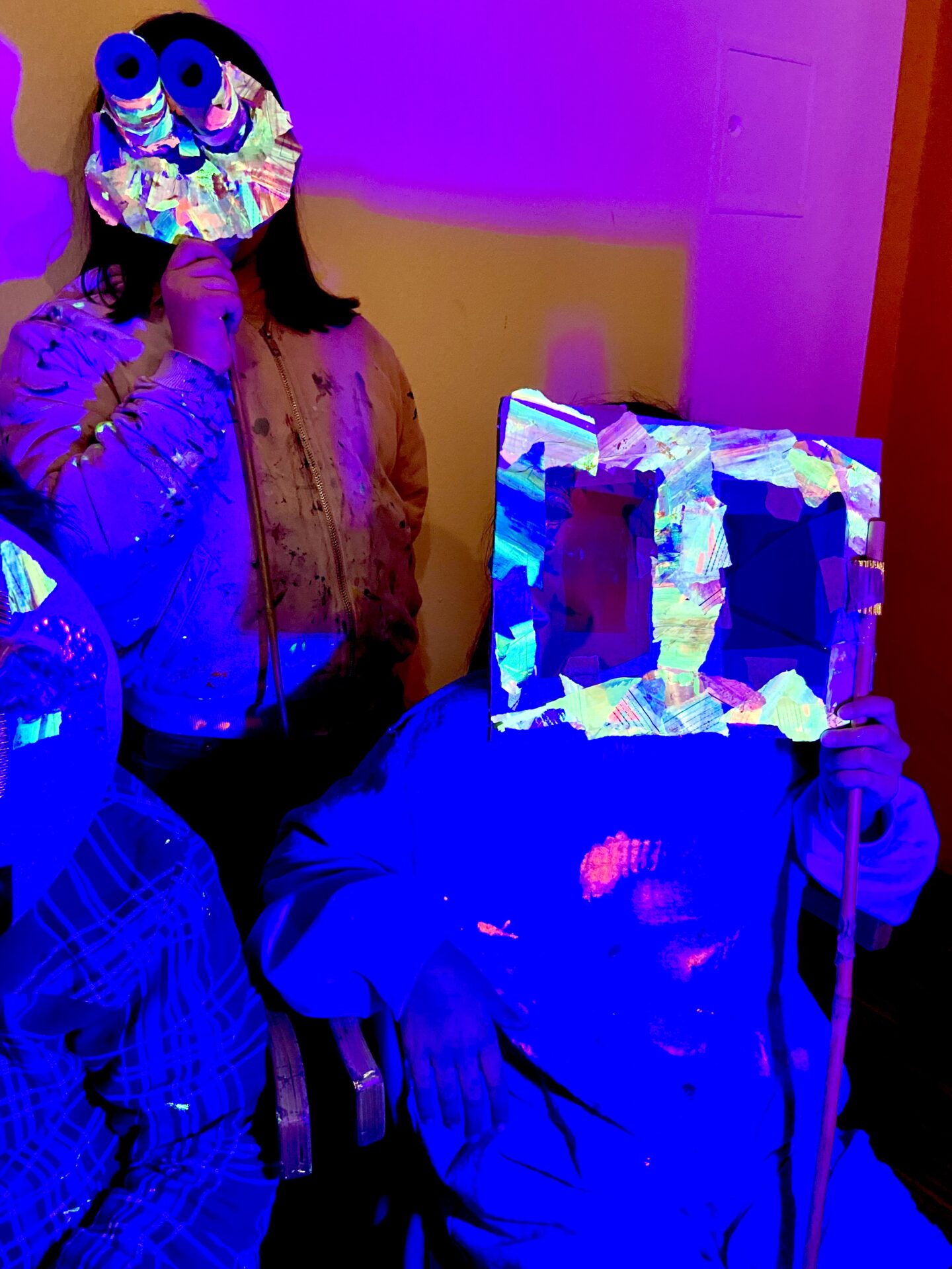 Drei Kinder halten sich bunte Masken vors Gesicht, die im Schwarzlicht leuchten.