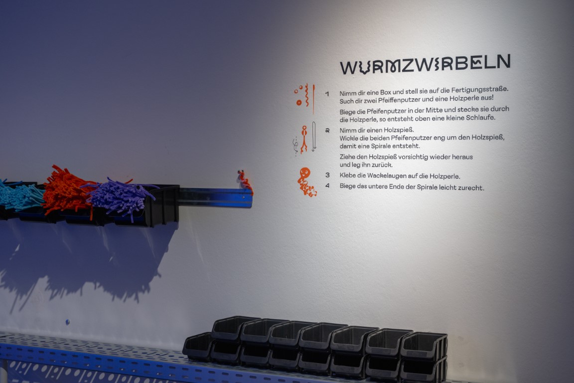 Zwei Leisten mit dunklen Boxen und Pfeifenputzern hängen an der Wand, daneben befindet sich eine Anleitung, wie daraus Wurmzwirbeln gebastelt werden sollen.