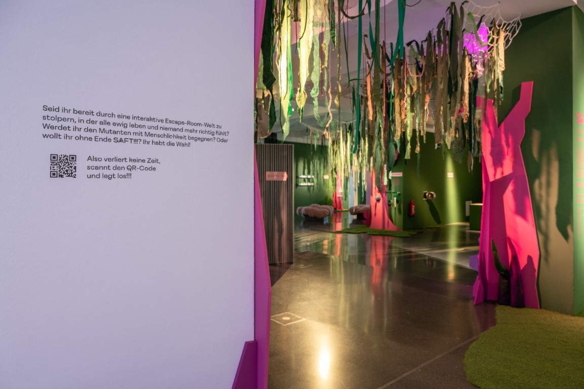 Im Hintergrund sind pinke und grüne Wände zu sehen, von der Decke hängen grüne Stoffbahnen.