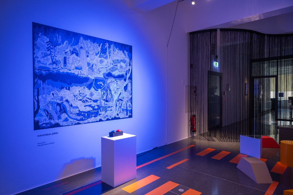 Blau beleuchtetes Bild an einer Wand vor dem ein weißer Sockel mit einem roten und einem blauen Buzzer drauf steht.