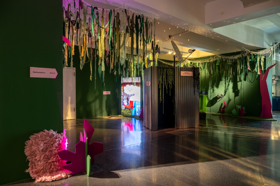Ausstellungsansicht der Ausstellung „Saft!!!“, im Zentrum steht eine dunkle Box mit offenem Eingang von dem Raken hängen.