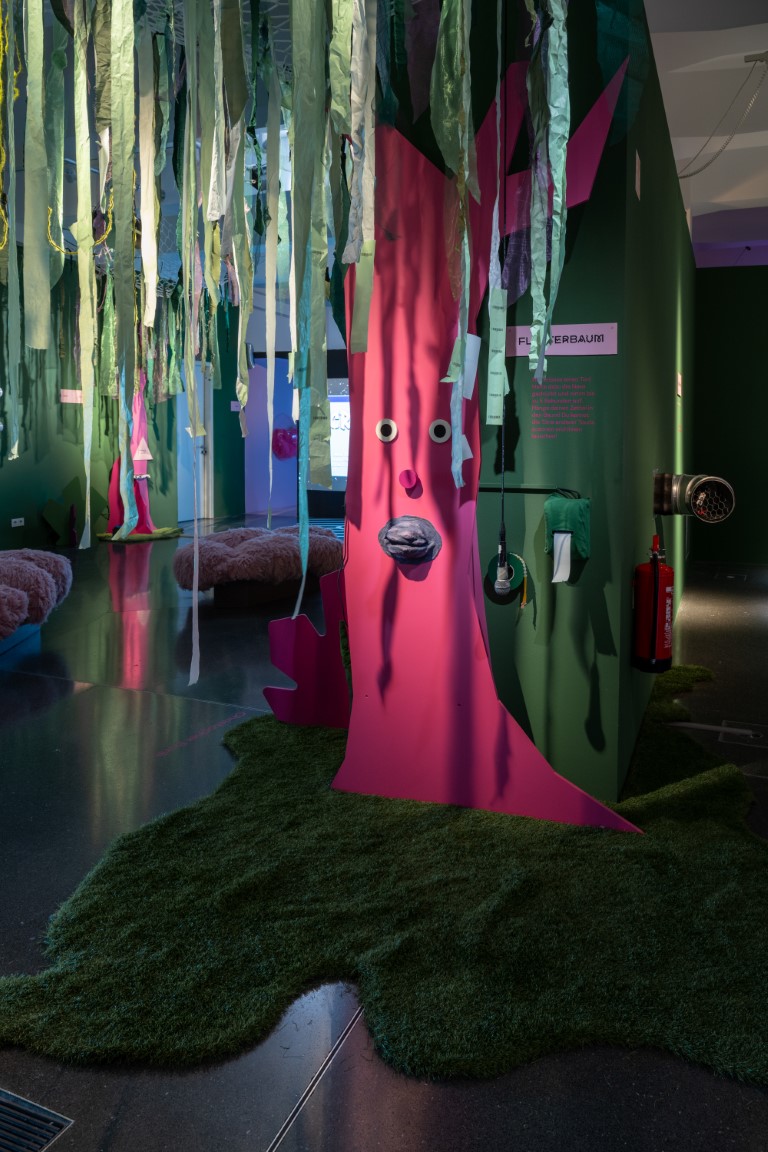 Vor einer grünen Wand steht ein pinker Baumstamm mit einem Gesicht auf einer Art Moosgrund, von der Decke hängen grüne Stoffstreifen.