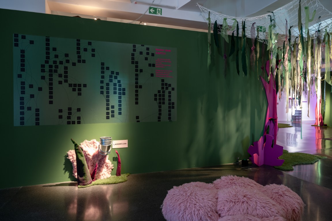 An einer grünen Wnad hängt ein Kunstwerk das aus vielen kleinen Feldern besteht die miteinander verbunden sind, darunter befindet sich ein Rohr das aus der Wand schaut, davor liegt ein großer, rosa flauschiges Kissen.