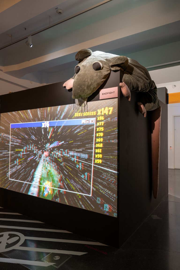 Auf einer großen schwarzen Fläche ist ein Spiel im Weltall projiziert, in der rechten oberen Ecke sitzt eine große Stoffratte.