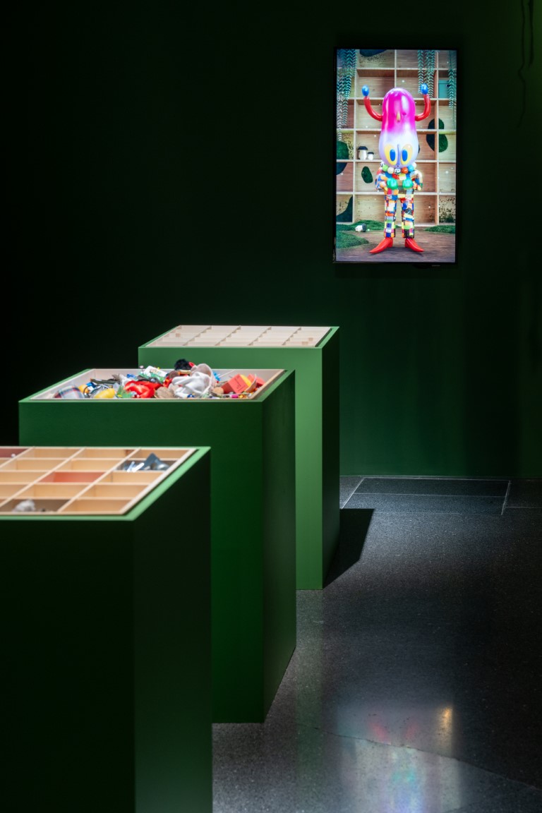 In drei grüne Sockeln sind Kisten mit verschiedenen Gegenständen eingelassen, im Hintergrund ist ein Bildschirm an der Wand.
