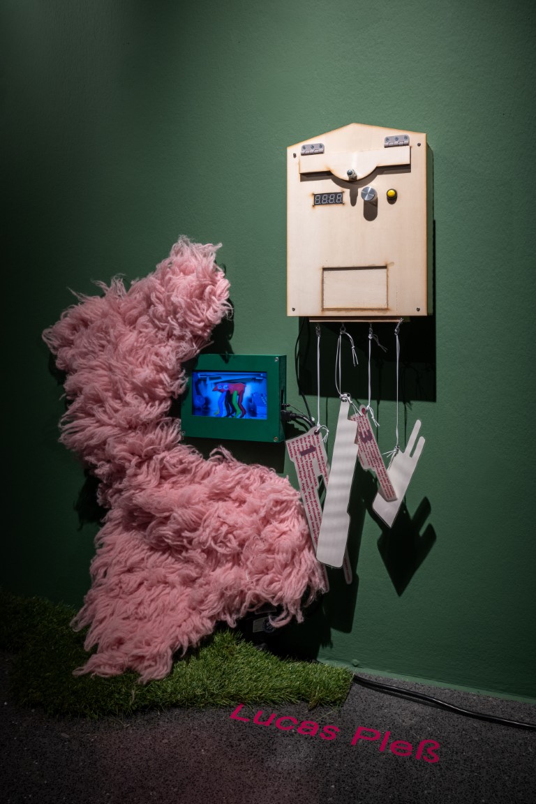 An einer grünen Wand hängt ein Holzkasten, links daneben befindet sich ein Bildschirm in einem grünen Rahmen, daneben ist rosa flauschiger Stoff an Wand und Boden.