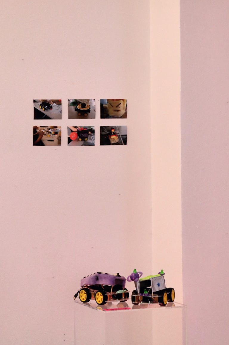 Im Hintergrund hängen Fotos an der Wand, im Vordergrund steht ein gebasteltes Auto auf einer Säule.