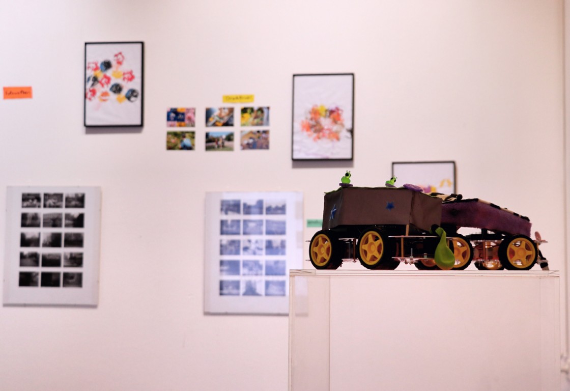 Im Hintergrund hängen Fotos an der Wand, im Vordergrund steht ein gebasteltes Auto auf einer Säule.