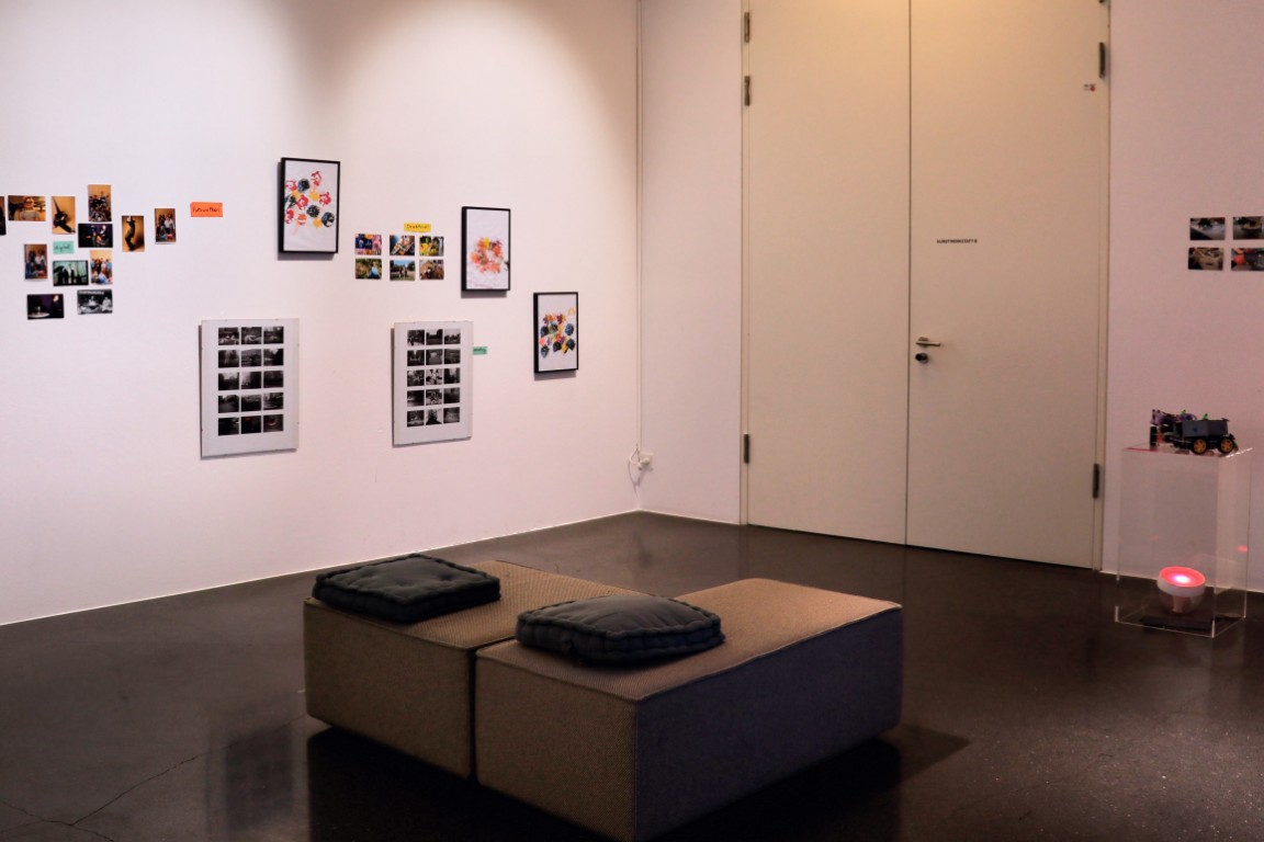 In der Mitte des Raumes stehen graue Sitzmöbel, im Hintergrund an der Wand hängen Fotos.