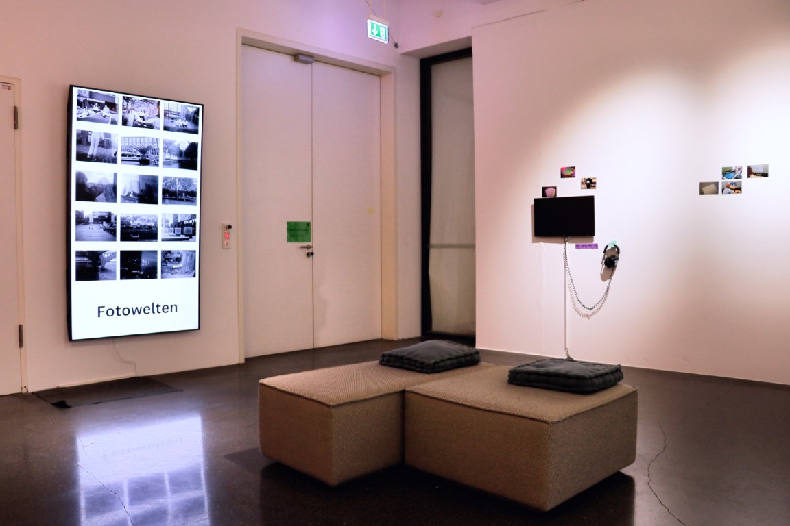 In der Mitte des Raumes stehen graue Sitzmöbel, im Hintergrund sind ein großer Bildschirm und ein Fernseher zu sehen.